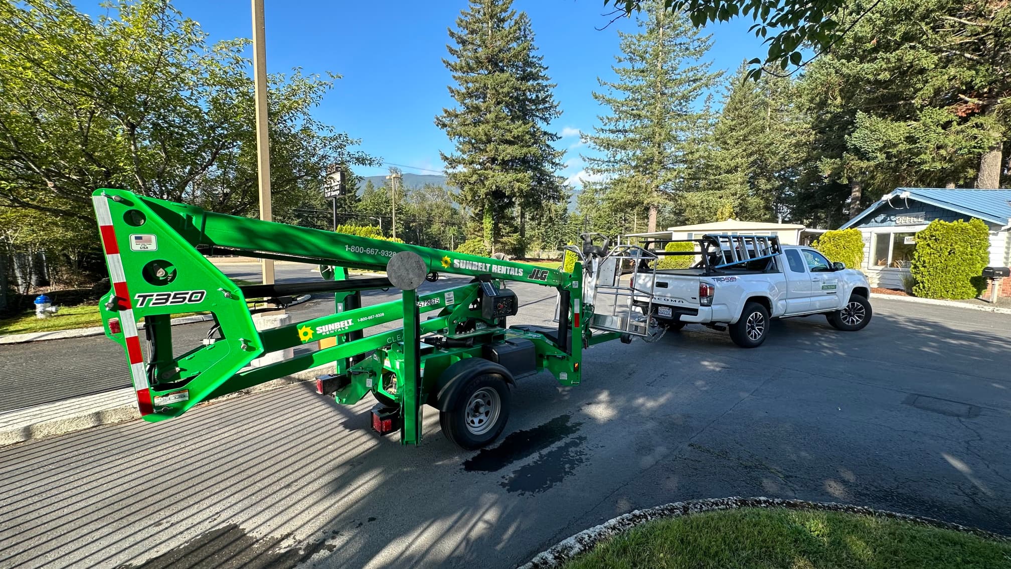 White Toyota Tacoma work truck with ladder racks parked next to a green Sunbelt Rentals T350 towable aerial lift, used for electrical installations and maintenance. The outdoor setting features trees, a blue sky, and a small office building, highlighting professional equipment for residential and commercial electrical projects.